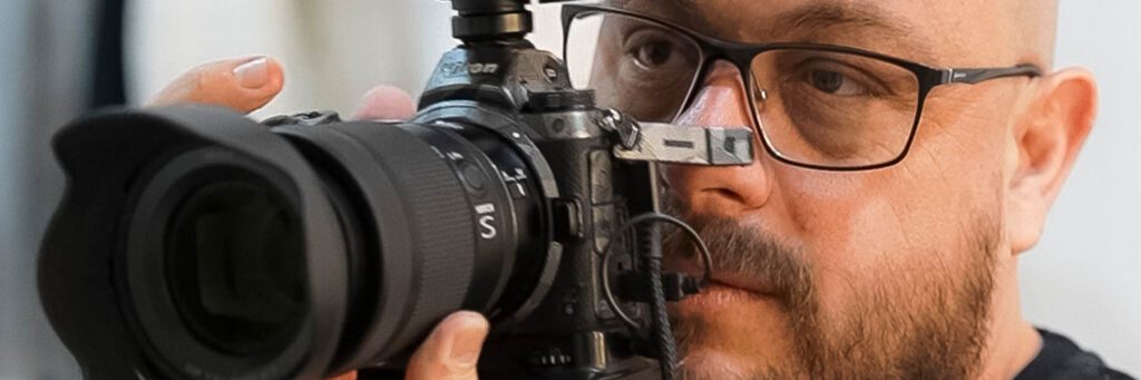 Retrato Corporativo - Alê Fotográfo - A imagem mostra um homem usando óculos e segurando uma câmera fotográfica profissional. Ele está olhando pelo visor da câmera, que está posicionada próxima ao seu rosto. A câmera tem uma lente grande e parece ser um modelo avançado. O homem tem barba e está concentrado na atividade de fotografar.